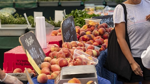 A bustling market scene features rows of fresh peaches displayed in open containers beside chalkboard signs that read 'organic'. A person carrying a black tote bag and wearing a grey T-shirt is partly visible on the right. The background includes stacks of greenery and more produce.