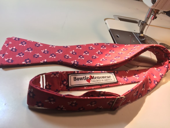 A red, floral-patterned bow tie lies adjacent to a sewing machine. The label on the bow tie reads 'Bowtie Menswear Tailored in America.'