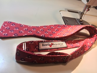 A red, floral-patterned bow tie lies adjacent to a sewing machine. The label on the bow tie reads 'Bowtie Menswear Tailored in America.'