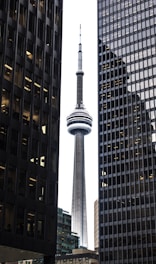 A tall tower with a cylindrical observation deck at the top is framed by two high-rise office buildings. The tower is highly prominent, with a sleek, modern design. The surrounding buildings have numerous windows, some illuminated, and create a symmetrical composition.