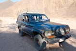 A rugged, dark blue Toyota SUV is parked on a sandy dirt road amidst a mountainous desert landscape. The vehicle appears weathered, with visible wear around the wheel arches and bumper. The sunlight casts shadows and highlights the texture of the rocky mountains in the background.