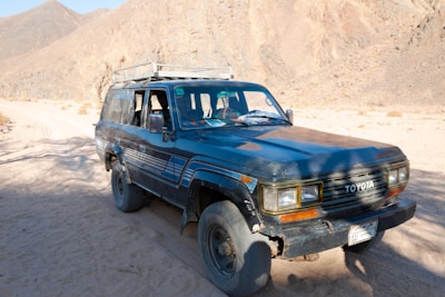A rugged, dark blue Toyota SUV is parked on a sandy dirt road amidst a mountainous desert landscape. The vehicle appears weathered, with visible wear around the wheel arches and bumper. The sunlight casts shadows and highlights the texture of the rocky mountains in the background.