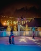 Two children wearing helmets are ice skating at an outdoor rink surrounded by festive lights. One child is using a penguin-shaped skate aid. Trees decorated with string lights are visible in the background, and there are cozy wooden structures under a misty, dark sky.