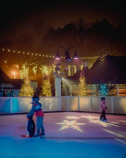 Two children wearing helmets are ice skating at an outdoor rink surrounded by festive lights. One child is using a penguin-shaped skate aid. Trees decorated with string lights are visible in the background, and there are cozy wooden structures under a misty, dark sky.