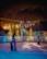 Two children wearing helmets are ice skating at an outdoor rink surrounded by festive lights. One child is using a penguin-shaped skate aid. Trees decorated with string lights are visible in the background, and there are cozy wooden structures under a misty, dark sky.