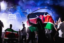 A vibrant concert scene with people holding flags against a backdrop of colorful lights and smoke. The logo 'Afro Bros' is prominently displayed in the background, and the atmosphere is dynamic and lively.