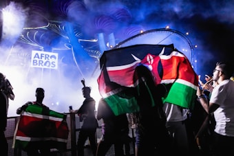 A vibrant concert scene with people holding flags against a backdrop of colorful lights and smoke. The logo 'Afro Bros' is prominently displayed in the background, and the atmosphere is dynamic and lively.