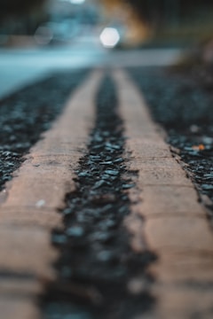 A close-up view of a road surface featuring two yellow parallel lines. The image focuses on the rough texture of the pavement with small rocks embedded within. The background is blurred, creating a sense of depth and an urban environment.