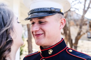 A person in a military uniform is smiling while looking at someone. The uniform features gold buttons and a distinctive emblem. In the background, there are trees and blurred structures, suggesting an outdoor setting.