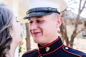 A person in a military uniform is smiling while looking at someone. The uniform features gold buttons and a distinctive emblem. In the background, there are trees and blurred structures, suggesting an outdoor setting.