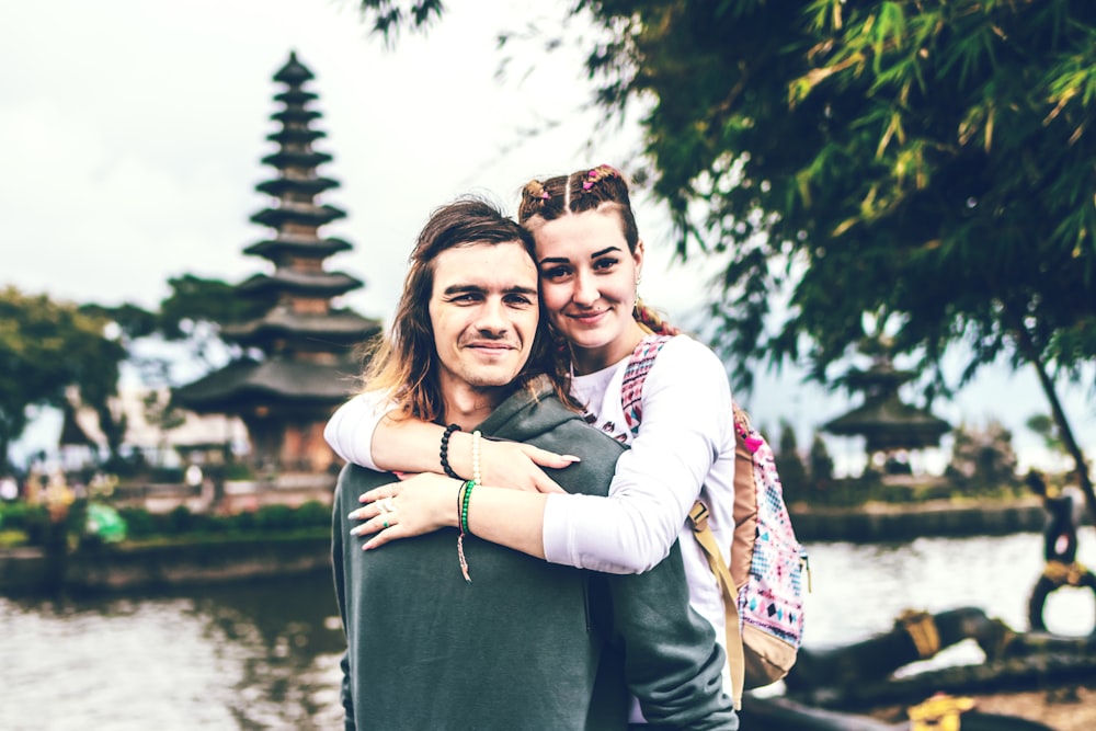 woman hugging man's back in front of body of water and tower