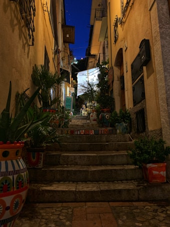 A narrow, dimly lit alleyway with stone steps leads up between two closely situated buildings. The alley is decorated with potted plants and colorful painted vases. Above, strands of lights hang, illuminating the upper part of the alley. A sign at the bottom of the stairs reads 'I &hearts; TAORMINA'. The sky is a deep blue, indicating early evening.