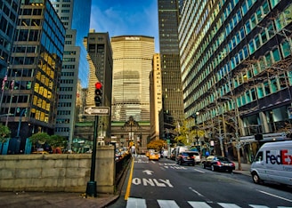 A bustling urban scene featuring tall skyscrapers lining both sides of the street, with various vehicles including cars and a FedEx delivery van on the road. The view includes a prominent building with 'MetLife' signage. Pedestrians and other urban elements such as traffic lights and signs are visible.