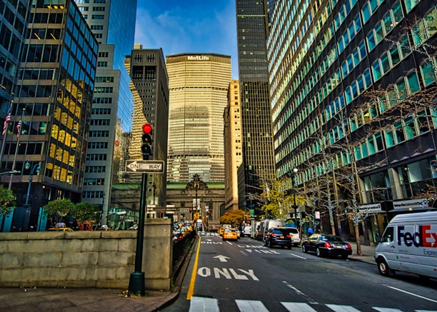 A bustling urban scene featuring tall skyscrapers lining both sides of the street, with various vehicles including cars and a FedEx delivery van on the road. The view includes a prominent building with 'MetLife' signage. Pedestrians and other urban elements such as traffic lights and signs are visible.