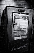 A weathered newspaper vending machine against a brick wall, featuring a publication titled 'Seattle Weekly'. The cover includes various headlines about labor practices and is marked with graffiti. The scene is in black and white, emphasizing the textures and shadows.