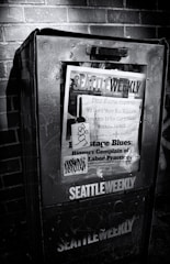 A weathered newspaper vending machine against a brick wall, featuring a publication titled 'Seattle Weekly'. The cover includes various headlines about labor practices and is marked with graffiti. The scene is in black and white, emphasizing the textures and shadows.