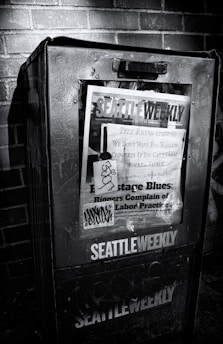 A weathered newspaper vending machine against a brick wall, featuring a publication titled 'Seattle Weekly'. The cover includes various headlines about labor practices and is marked with graffiti. The scene is in black and white, emphasizing the textures and shadows.