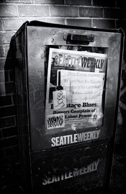 A weathered newspaper vending machine against a brick wall, featuring a publication titled 'Seattle Weekly'. The cover includes various headlines about labor practices and is marked with graffiti. The scene is in black and white, emphasizing the textures and shadows.