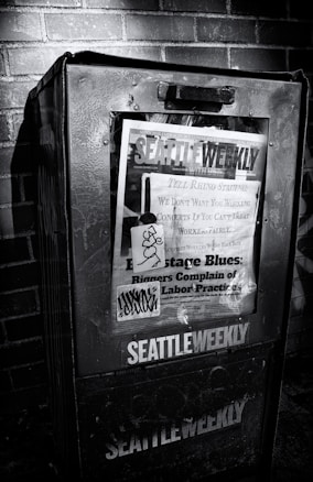 A weathered newspaper vending machine against a brick wall, featuring a publication titled 'Seattle Weekly'. The cover includes various headlines about labor practices and is marked with graffiti. The scene is in black and white, emphasizing the textures and shadows.
