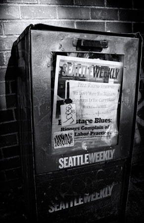 A weathered newspaper vending machine against a brick wall, featuring a publication titled 'Seattle Weekly'. The cover includes various headlines about labor practices and is marked with graffiti. The scene is in black and white, emphasizing the textures and shadows.