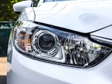 A close-up view of a car headlight with intricate design details, featuring clear lenses and chrome accents. The surrounding body of the car is painted white, reflecting light and capturing the sleek, modern design.