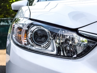 A close-up view of a car headlight with intricate design details, featuring clear lenses and chrome accents. The surrounding body of the car is painted white, reflecting light and capturing the sleek, modern design.