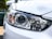 A close-up view of a car headlight with intricate design details, featuring clear lenses and chrome accents. The surrounding body of the car is painted white, reflecting light and capturing the sleek, modern design.