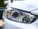 A close-up view of a car headlight with intricate design details, featuring clear lenses and chrome accents. The surrounding body of the car is painted white, reflecting light and capturing the sleek, modern design.