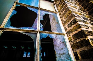 A shattered window with large, sharp fragments of glass in various shapes and sizes. The window is set in an old brick wall, showing signs of wear and age. Light reflects off the broken glass, creating a contrast between the dark interior and the brighter exterior.