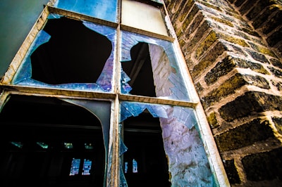 A shattered window with large, sharp fragments of glass in various shapes and sizes. The window is set in an old brick wall, showing signs of wear and age. Light reflects off the broken glass, creating a contrast between the dark interior and the brighter exterior.