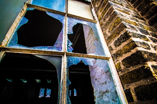 A shattered window with large, sharp fragments of glass in various shapes and sizes. The window is set in an old brick wall, showing signs of wear and age. Light reflects off the broken glass, creating a contrast between the dark interior and the brighter exterior.