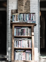 A wooden bookshelf filled with a variety of books, located against a white brick wall. Above the bookshelf, there is a handwritten sign listing various categories such as art, poetry, and health. The setting appears cozy and perhaps within a rustic or vintage-style room.