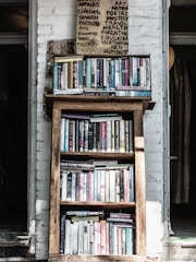 A wooden bookshelf filled with a variety of books, located against a white brick wall. Above the bookshelf, there is a handwritten sign listing various categories such as art, poetry, and health. The setting appears cozy and perhaps within a rustic or vintage-style room.
