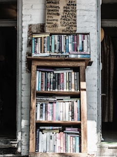 A wooden bookshelf filled with a variety of books, located against a white brick wall. Above the bookshelf, there is a handwritten sign listing various categories such as art, poetry, and health. The setting appears cozy and perhaps within a rustic or vintage-style room.