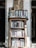 A wooden bookshelf filled with a variety of books, located against a white brick wall. Above the bookshelf, there is a handwritten sign listing various categories such as art, poetry, and health. The setting appears cozy and perhaps within a rustic or vintage-style room.