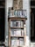 A wooden bookshelf filled with a variety of books, located against a white brick wall. Above the bookshelf, there is a handwritten sign listing various categories such as art, poetry, and health. The setting appears cozy and perhaps within a rustic or vintage-style room.