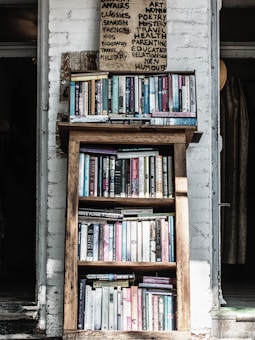 A wooden bookshelf filled with a variety of books, located against a white brick wall. Above the bookshelf, there is a handwritten sign listing various categories such as art, poetry, and health. The setting appears cozy and perhaps within a rustic or vintage-style room.