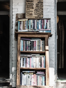 A wooden bookshelf filled with a variety of books, located against a white brick wall. Above the bookshelf, there is a handwritten sign listing various categories such as art, poetry, and health. The setting appears cozy and perhaps within a rustic or vintage-style room.