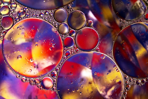 A vibrant composition of variously sized oil droplets suspended on a colorful surface, creating a stunning abstract pattern. The droplets are round and reflect light, showcasing a mix of reds, yellows, blues, and purples. The background displays a swirling mélange of colors, adding depth to the image.