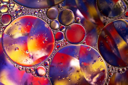 A vibrant composition of variously sized oil droplets suspended on a colorful surface, creating a stunning abstract pattern. The droplets are round and reflect light, showcasing a mix of reds, yellows, blues, and purples. The background displays a swirling mélange of colors, adding depth to the image.