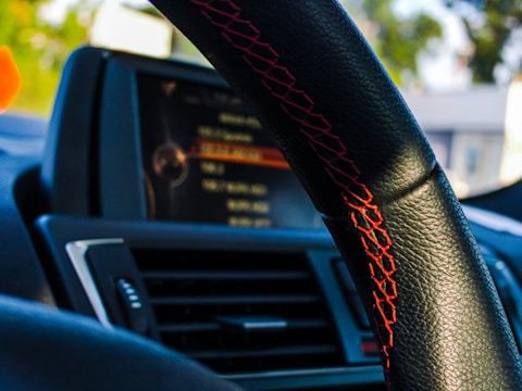 A close-up view of a car interior focusing on the steering wheel with red stitching. The background shows a blurred view of a car's dashboard display, with visible buttons and vents, suggesting a modern vehicle design.