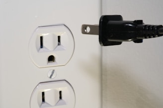A white electrical outlet with two sockets is positioned on the left side. Near it is a black electrical plug with two prongs oriented towards the outlet, suggesting a connection is about to be made.