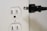 A white electrical outlet with two sockets is positioned on the left side. Near it is a black electrical plug with two prongs oriented towards the outlet, suggesting a connection is about to be made.