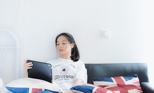 A young person with a relaxed expression sits on a sofa, holding a tablet. The background is a plain white wall, and colorful pillows with the Union Jack design flank the person.