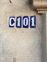 A textured beige wall features blue and white ceramic tiles displaying the characters C101. The tiles are rectangular and have a glossy finish, contrasting with the rough surface of the wall.