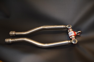 A pair of metallic suspension arms with rod ends, featuring a red center component for adjustment. The arms are made of shiny metal and have a smooth finish, resting on a dark background.