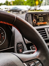 The interior of a car with a focus on the steering wheel, dashboard, and digital display. The digital display is showing a list of radio stations with the current selection highlighted. The start-stop engine button is visible near the steering wheel, which has a black and red design.