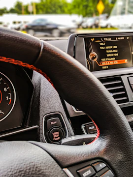 The interior of a car with a focus on the steering wheel, dashboard, and digital display. The digital display is showing a list of radio stations with the current selection highlighted. The start-stop engine button is visible near the steering wheel, which has a black and red design.
