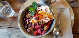 A bowl filled with a colorful assortment of fresh fruits and granola, topped with a dollop of yogurt and garnished with a sprig of mint. Slices of orange, chunks of watermelon, and various berries are visible, creating a vibrant presentation. A glass of water and a metal mug are placed beside the bowl on a wooden table, accompanied by a spoon and napkin.