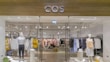 A clothing store display featuring mannequins dressed in casual attire, such as striped and checked shirts and skirts. The store interior is organized with neatly arranged racks of various clothing items, including shirts, blouses, and jackets in a range of colors such as yellow, white, pink, black, and blue. The entrance has glass doors, and the store name is prominently displayed above in bold letters. The lighting is bright, highlighting the clean, modern aesthetic of the space.
